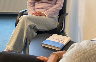 Teilansicht einer Person, die sitzend gegenüber einer anderen Person sitzt. Auf einem Beistelltisch liegt ein Buch aus der Psychologie.