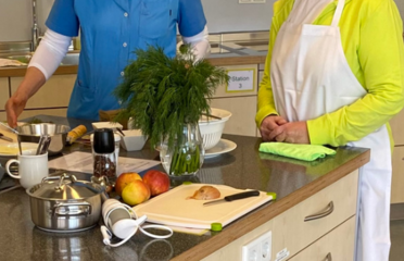 Zwei Personen in der Lehrküche, stehend an der Arbeitsfläche. Diverse Zutaten und Küchengeräte im Vordergrund.