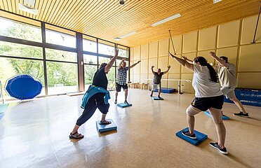 Gruppe von Personen, die im Kreis stehen in einer Bewegungshalle mit großer Fensterfront. Alle strecken beide Arme Richtung Decke und stehen in Schrittstellung mit dem vorderen Bein auf einem Balancekissen.