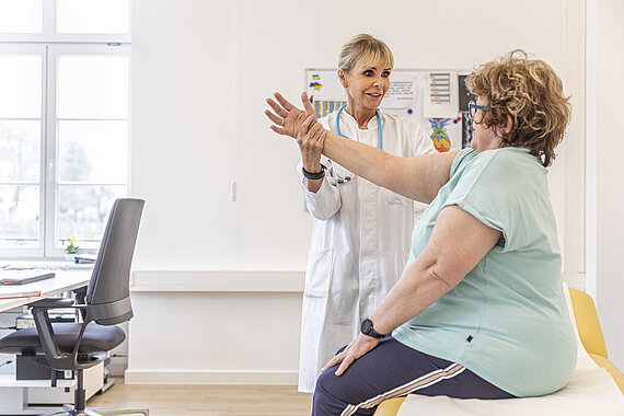 Ärztin bei einen Untersuchung einer Patientin, die auf einer Behandlungsliege in einem Arztzimmer sitzt. Die Ärztin hält den gestreckten Arm der Patientin auf Schulterhöhe hoch.