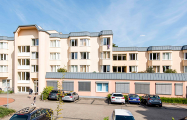 Gebäudebild vom Brunnenhof. Das Bettenhaus hat drei Etagen und besteht aus versetzten Gebäudeteilen mit einem Parkplatz davor.